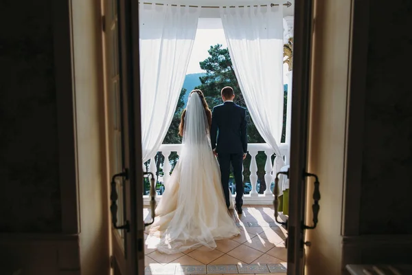 Jovem casal apaixonado por recém-casados de pé na varanda branca e de mãos dadas, vista traseira. A noiva em um belo vestido branco com cabelo comprido e um véu abraça o noivo em um terno clássico preto . — Fotografia de Stock