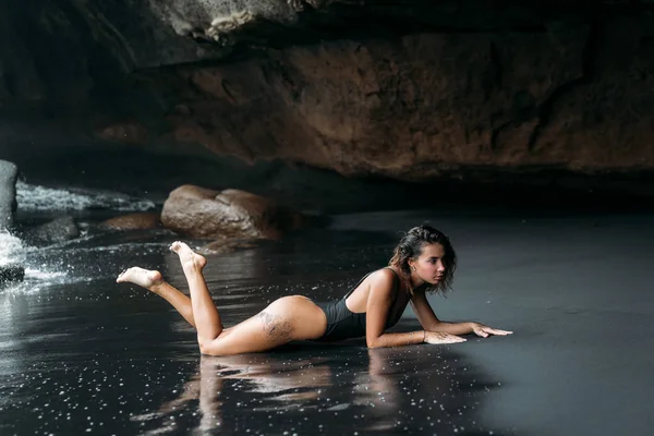 Красива дівчина з сексуальним тілом в чорному купальнику лежить на пляжі з чорним вулканічним піском. Молода модель з кучерявими волоссям, танірована шкіра позує і розслабляється в подорожі . — стокове фото