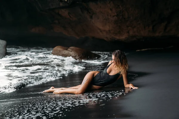 Красивая девушка с сексуальным телом в черном купальнике лежит на пляже с черным вулканическим песком. Молодая модель с вьющимися волосами, загорелой кожей позирует и расслабляется в путешествии . — стоковое фото