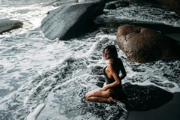 Von oben und von hinten, Porträt eines schönen Mädchens mit einem sexy Körper in einem schwarzen Badeanzug am Strand mit schwarzem Vulkansand. schönes junges Model mit sportlichem Körper und gebräunter Haut posiert auf einem — Stockfoto