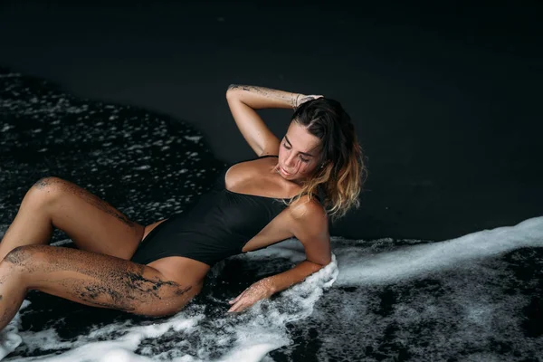 A chic girl with a sexy figure lies in a swimsuit on the beach with black volcanic sand. Model with a tanned body sunbathing on rest. — Stock Photo, Image