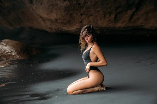 Uma garota incrível com uma figura sexy se senta em um maiô na praia com areia vulcânica preta. Modelo com um corpo bronzeado banhos de sol em férias em Bali . — Fotografia de Stock