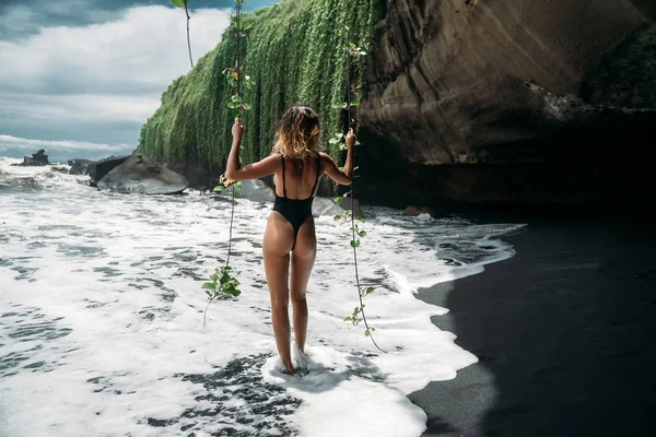 Rückansicht, ein Mädchen mit einem sexy Körper und einem Esel in einem schwarzen Badeanzug am Strand mit schwarzem Vulkansand. schönes junges Model mit sportlichem Körper und gebräunter Haut posiert am Meeresstrand — Stockfoto