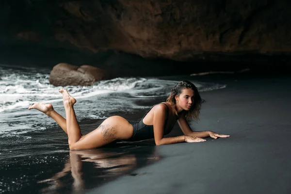 Belle fille au corps sexy en maillot de bain noir couché sur la plage avec du sable volcanique noir. Jeune modèle aux cheveux bouclés, peau bronzée posant et relaxant dans le voyage . — Photo