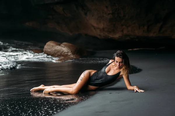 Menina bonita com corpo sexy em maiô preto deitado na praia com areia vulcânica preta. Modelo jovem com cabelos encaracolados, pele bronzeada posando e relaxando na viagem . — Fotografia de Stock