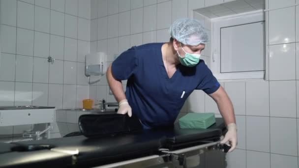 De arts in het ziekenhuis past de chirurgische tabel. De chirurg bereidt voor chirurgie, een tabel en medische apparatuur in de kliniek. Een medische professional in steriele kleding werkt in een kamer met — Stockvideo