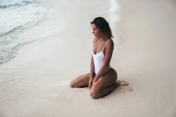 Гонжерообразная модель в белом купальнике с красивым спортивным телом лежит и позирует на белом песчаном пляже. Загорелая молодая женщина с вьющимися волосами отдыхает и загорает. Очаровательная модель у моря . — стоковое фото