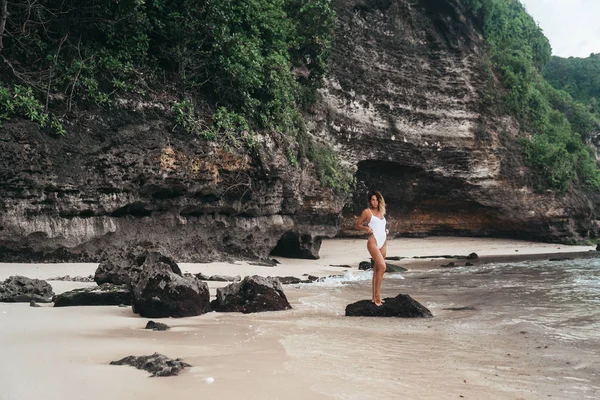 Erstaunliche Mädchen in einem weißen Badeanzug mit einem schönen Sportkörper gehen und posieren an einem weißen Sandstrand. Eine braun gebrannte junge Frau mit lockigem Haar ruht und sonnt sich. Charmantes Model posiert in der Nähe der — Stockfoto