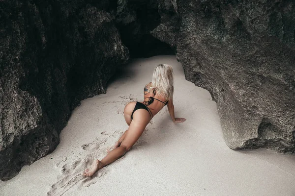美しいボディとの入れ墨を持つ少女は、彼女の膝の上、砂浜に沿ってゾッし、彼女のお尻を示しています。白いビーチで休んで黒の水着でセクシーなモデル. — ストック写真