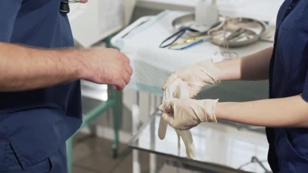 Se está preparando un primer equipo de médicos para la operación. El asistente de enfermería ayuda al cirujano a ponerse guantes blancos. Herramientas médicas en segundo plano. Concepto de salud, trabajo en el hospital. Equipos — Vídeos de Stock