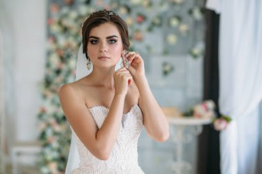Tatlı bir genç gelin düğün günü gidiyor ve onun kulağına küpe üzerinde koyarak. Saç ve makyaj tatil için hazırlanan güzel esmer kız. Saç modeli içinde diadem. Pahalı şık elbise