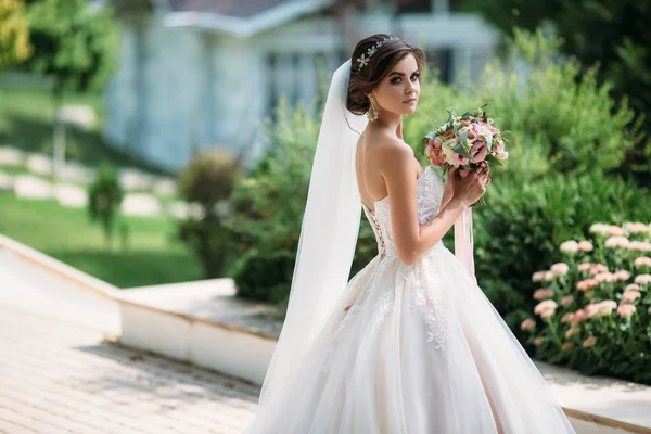 Schöne Luxus-Braut mit Braut Make-up und Frisur in der Ehe. Porträt des Models in einem wunderschönen weißen Hochzeitskleid mit einem Blumenstrauß in ihren Händen. Konzept der Familie. — Stockfoto