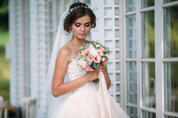 Retrato de noiva bonita com flores de buquê em vestido de luxo branco. Modelo lindo com maquiagem nupcial e penteado com um diadema em seu cabelo, brincos de diamante no ouvido. Casamento e florística — Fotografia de Stock