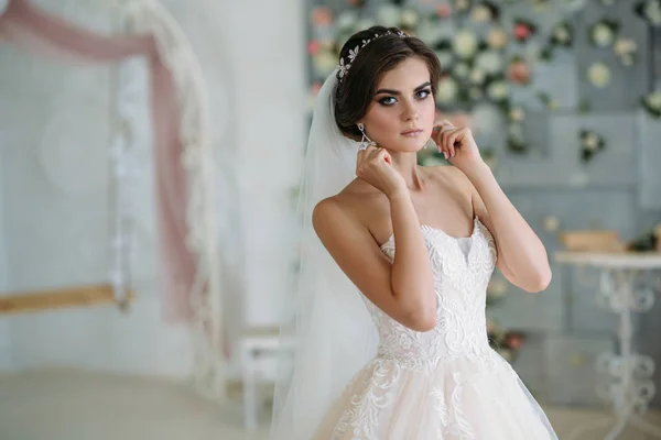 Uma jovem noiva doce no dia do casamento está indo e colocando brincos em seu ouvido. Menina morena bonita com cabelo e maquiagem preparada para o feriado. Diadem em penteado. Vestido elegante caro — Fotografia de Stock