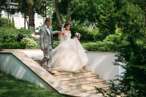 Um casal feliz a abraçar-se e a passear no parque. A noiva em um terno preto com um laço e uma noiva em um vestido de noiva branco bonito com um buquê de flores em suas mãos. Mulher bonita com — Fotografia de Stock