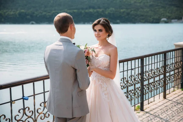 Bella sposa con i capelli lunghi in abito bianco abbracciando sposo e sorridente carino. Affascinante coppia di sposi il giorno del matrimonio, lago sullo sfondo. Una famiglia felice all'aperto nel parco . — Foto Stock