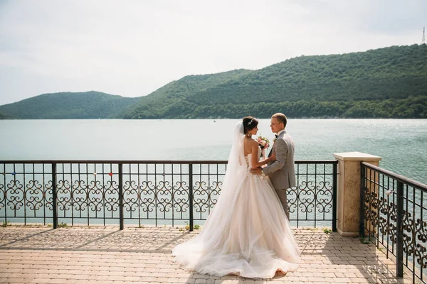 美しい結婚式のカップル、花嫁と花婿の湖の背景に手を繋いでいます。長い白いドレス、灰色の背広の男性にかわいい女の子。背景に山の風景。家族の概念. — ストック写真