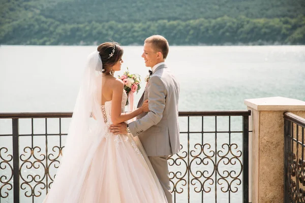 Vackra bröllopsparet, bruden och brudgummen håller händerna på sjön bakgrund. Söt flicka i lång vit klänning, män i grå kostym. landskap i bakgrunden. Begreppet familj. — Stockfoto