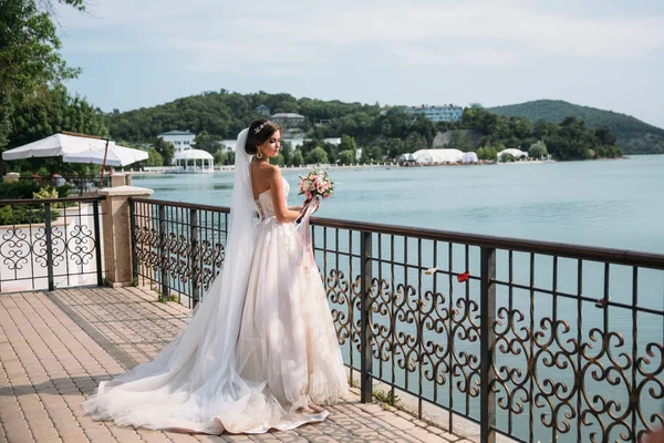 Bela noiva jovem em vestido de noiva branco com cabelos longos, flores de buquê em suas mãos stands, lago no fundo . — Fotografia de Stock