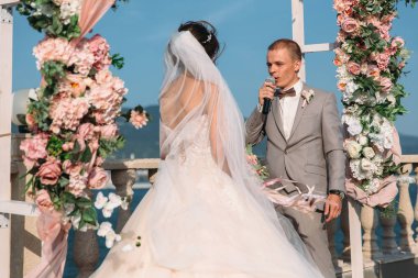 Çekici damat ve geline düğün günü ceremory kemer ve arka plan standları birlikte gölde. Güzel yeni evliler, beyaz elbise ve uzun tüyleri, siyah takım elbise giyen adamlar genç kadın. Mutlu bir aile