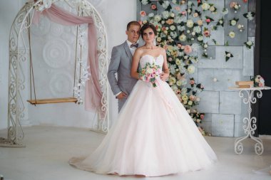 Gelin Damat hugs ve bir buket çiçek onun elinde tutar. Güzel birkaç hafif bir stüdyoda poz bir düğün gününde yeni evliler. Bir erkekte bir lüks gri takım elbise, beyaz bir gömlek ve