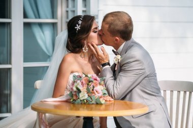 öpüşme ve sarılma masada oturan yeni evliler. Gelin ve damat birbirine bakarak. Bir adam bir gri takım elbise, beyaz bir gömlek ve bir papyon. Beyaz düğün makyajlı genç kız