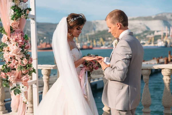 Coppia in anelli di scambio di arco nuziale con lago su sfondo, la sposa con bei capelli lunghi e sposo in costume nero guardano all'un l'altro in giorno nuziale. Concetto di amore e famiglia — Foto Stock