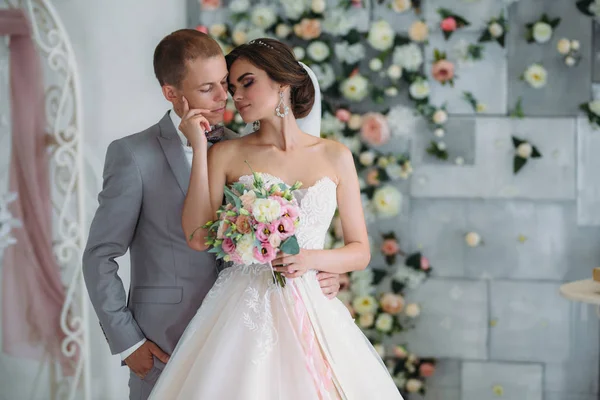 Hermosa pareja de boda abrazándose en estudio luminoso. El novio en un traje gris de negocios, una camisa blanca con pajarita y un ojal, una novia en un vestido de lujo con un velo y un ramo de flores — Foto de Stock