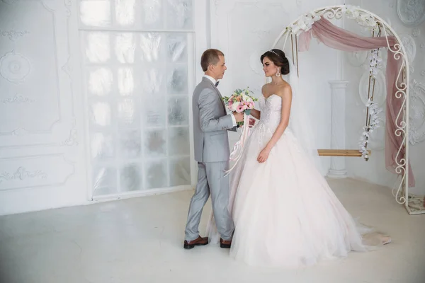 Umarmt die Braut den Bräutigam und hält einen Blumenstrauß in ihren Händen. ein schönes Brautpaar an einem Hochzeitstag, das in einem hellen Studio posiert. ein Mann in einem luxuriösen grauen Business-Anzug, einem weißen Hemd und — Stockfoto