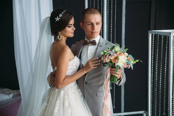 Umarmt die Braut den Bräutigam und hält einen Blumenstrauß in ihren Händen. ein schönes Brautpaar an einem Hochzeitstag, das in einem hellen Studio posiert. ein Mann in einem luxuriösen grauen Business-Anzug, einem weißen Hemd — Stockfoto