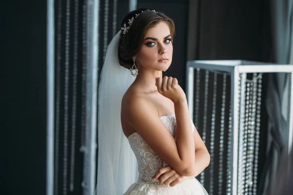 Preparação da noiva em um dia de casamento. Menina morena bonita em um vestido de luxo branco, com brincos, maquiagem e penteado posando em um estúdio escuro. Conceito de casamento e família . — Fotografia de Stock