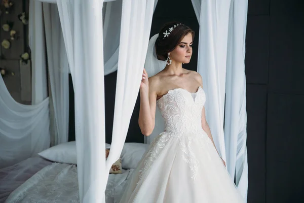 Preparação da noiva em um dia de casamento. Menina morena bonita em um vestido de luxo branco, com brincos, maquiagem e penteado posando em um estúdio escuro perto da cama e esperando o noivo. Conceito de — Fotografia de Stock