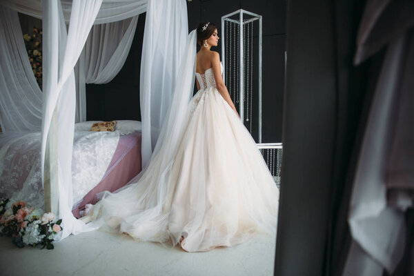preparation of the bride on a wedding day. Beautiful brunette girl in a white luxury dress, with earrings, make-up and hairdo posing in a dark studio near the bed and waiting for the groom. Concept of
