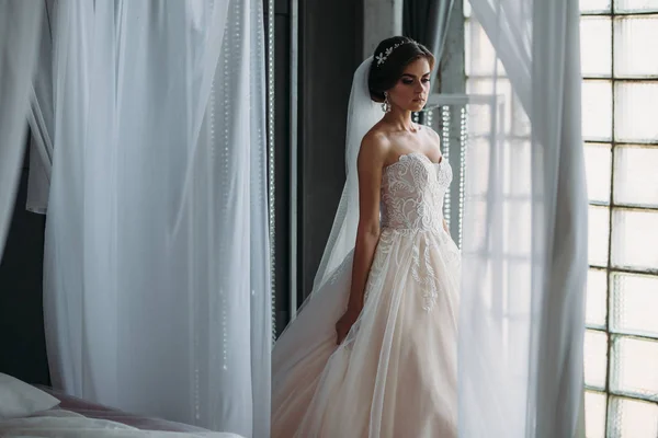 Preparation of the bride on a wedding day. Beautiful brunette girl in a white luxury dress, with earrings, make-up and hairdo posing in a dark studio. Concept of marriage and family. — Stock Photo, Image