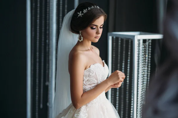 Vorbereitung der Braut auf einen Hochzeitstag. schöne brünette Mädchen in einem weißen Luxuskleid, mit Ohrringen, Make-up und Frisur posiert in einem dunklen Studio. Konzept von Ehe und Familie. — Stockfoto