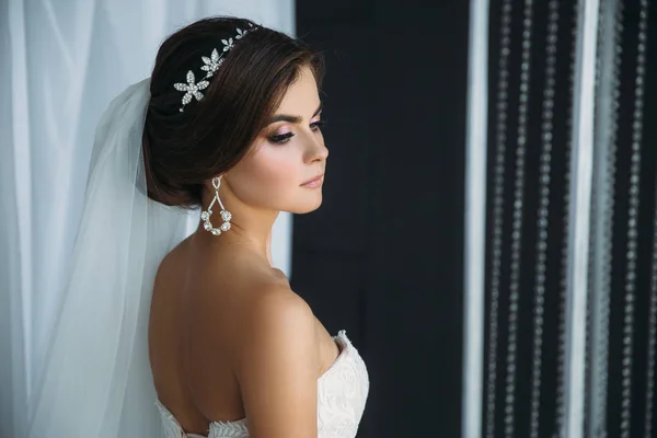 Preparação da noiva em um dia de casamento. Menina morena bonita em um vestido de luxo branco, com brincos, maquiagem e penteado posando em um estúdio escuro. Conceito de casamento e família . — Fotografia de Stock