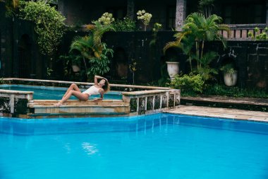 Yakınındaki bir havuz mavi su ile güneşlenme bikinili güzel kız. Seksi model spor şekil ile istirahat ediyor. Hafta sonları bir mayo kıvırcık saçlı genç kadın.
