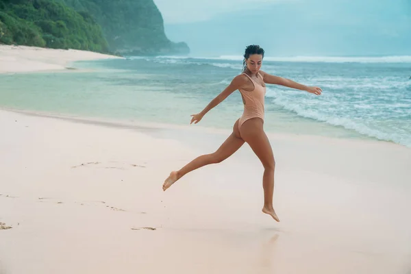Брюнетка с вьющимися волосами в бежевом купальнике занимается спортом на пляже с белым песком возле океана на отдыхе. Красивая модель с сексуальным телом загорает . — стоковое фото