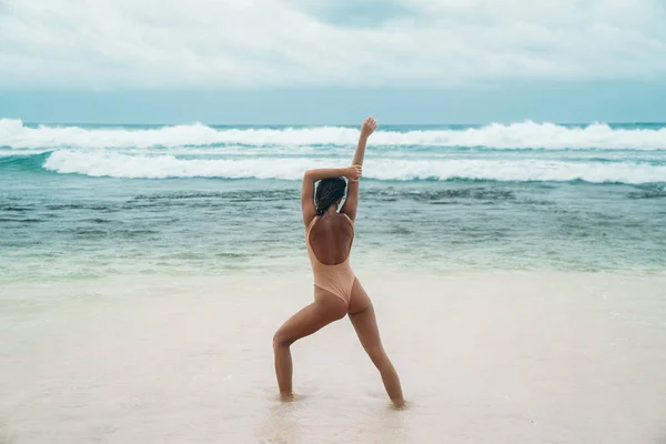 Brunette dívka s kudrnatými vlasy v béžových plavkách se zabývá sportovní na pláži s bílým pískem u moře na dovolené. Krásný model s sexy tělo je opalování. — Stock fotografie