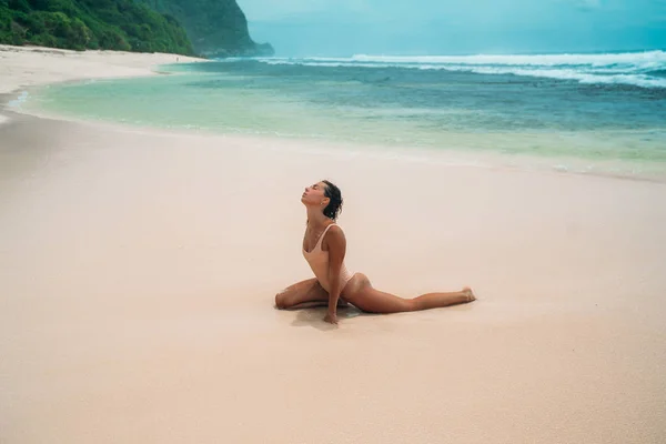 Bruna ragazza con i capelli ricci in costume da bagno beige è impegnata nello sport sulla spiaggia con sabbia bianca vicino all'oceano in vacanza. Un bel modello con un corpo sexy sta prendendo il sole . — Foto Stock