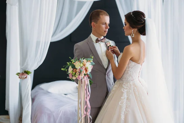 La novia en un lujoso vestido blanco con un velo se pone un novio boutonniere en un traje gris. Pareja de boda en el día de la boda de vestir. Preparación de los recién casados . — Foto de Stock