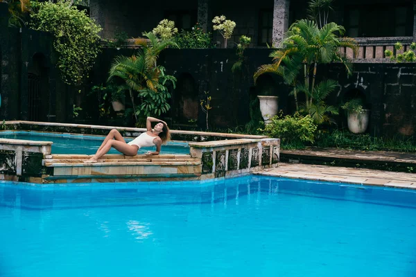 Hermosa chica en bikini tomando el sol cerca de una piscina con agua azul. Modelo sexy con una figura deportiva está descansando. Mujer joven con el pelo rizado en traje de baño en el fin de semana . — Foto de Stock