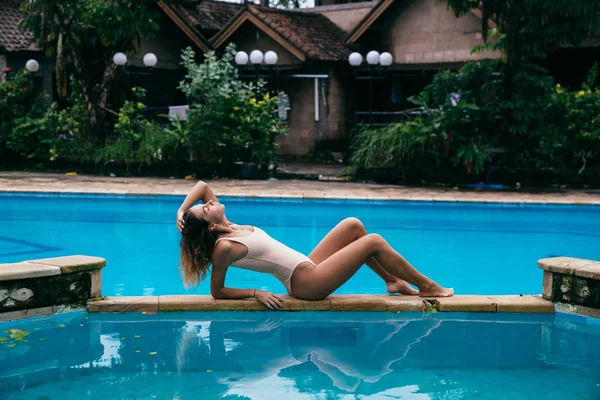 Beautiful girl in a bikini sunbathing and lies near a swimming pool with blue water. Sexy model with a sports figure is resting. Young woman with curly hair in a swimsuit at the weekend. — Stock Photo, Image