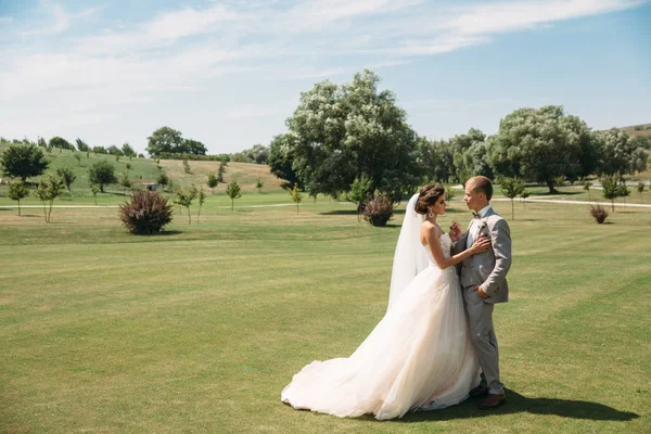 Gli sposi stanno camminando lungo il campo da golf con erba verde. Lo sposo tiene la mano delle spose. Bella coppia il giorno del matrimonio per una passeggiata. Ragazza in abito bianco lungo di lusso con velo, uomo in — Foto Stock