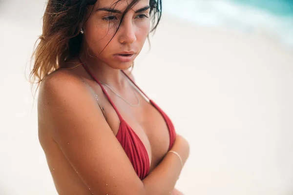 Portrait of a sexy tanned girl on the beach. A gorgeous model with a tight-fitting sports figure in a red bikini swimsuit. Young woman on vacation posing on white sand on the island. — Stock Photo, Image