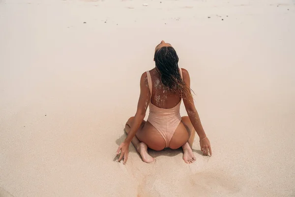 Vista posteriore, ragazza sexy in costume da bagno è impegnata in acqua yoga sulla spiaggia. Giovane donna con i capelli ricci posa e riposo vicino all'oceano. Bellissimo corpo sportivo di un turista sull'isola . — Foto Stock
