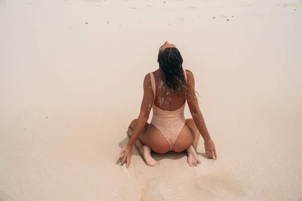 Rear view, sexy girl in a swimsuit is engaged in aqua yoga on the beach. Young woman with curly hair posing and resting near the ocean. Beautiful sports body of a tourist on the island. — Stock Photo, Image