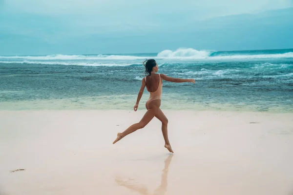 Brünettes Mädchen mit lockigem Haar im beigen Badeanzug treibt im Urlaub Sport am Strand mit weißem Sand am Meer. ein schönes Model mit einem sexy Körper sonnt sich. — Stockfoto