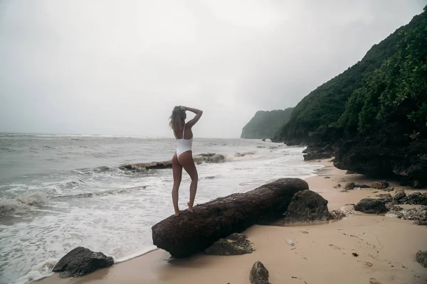 섹시 한 몸매와 아름 다운 엉덩이, 섹시 한 여자 다시 보기. 흰색 수영복 해변에 바위 근처에 매력적인 여자. 놀라운 볼 수 있는 섬에 여름 휴가에 포즈 모델. — 스톡 사진
