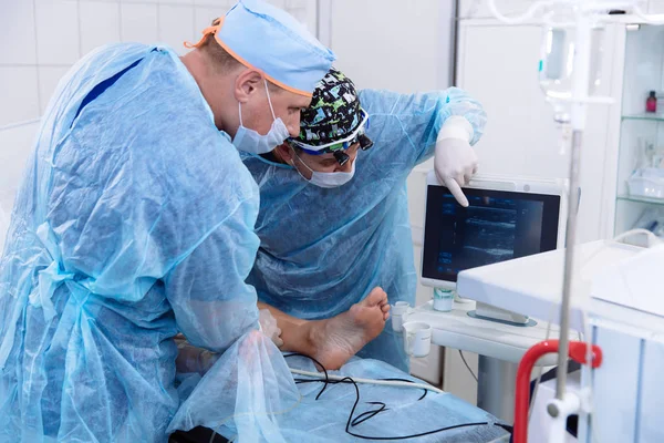 Un team di medici in abiti medici sterili eseguire un intervento chirurgico in una sala operatoria. Il chirurgo mostra l'apparecchiatura diagnostica e comunica con l'assistente . — Foto Stock